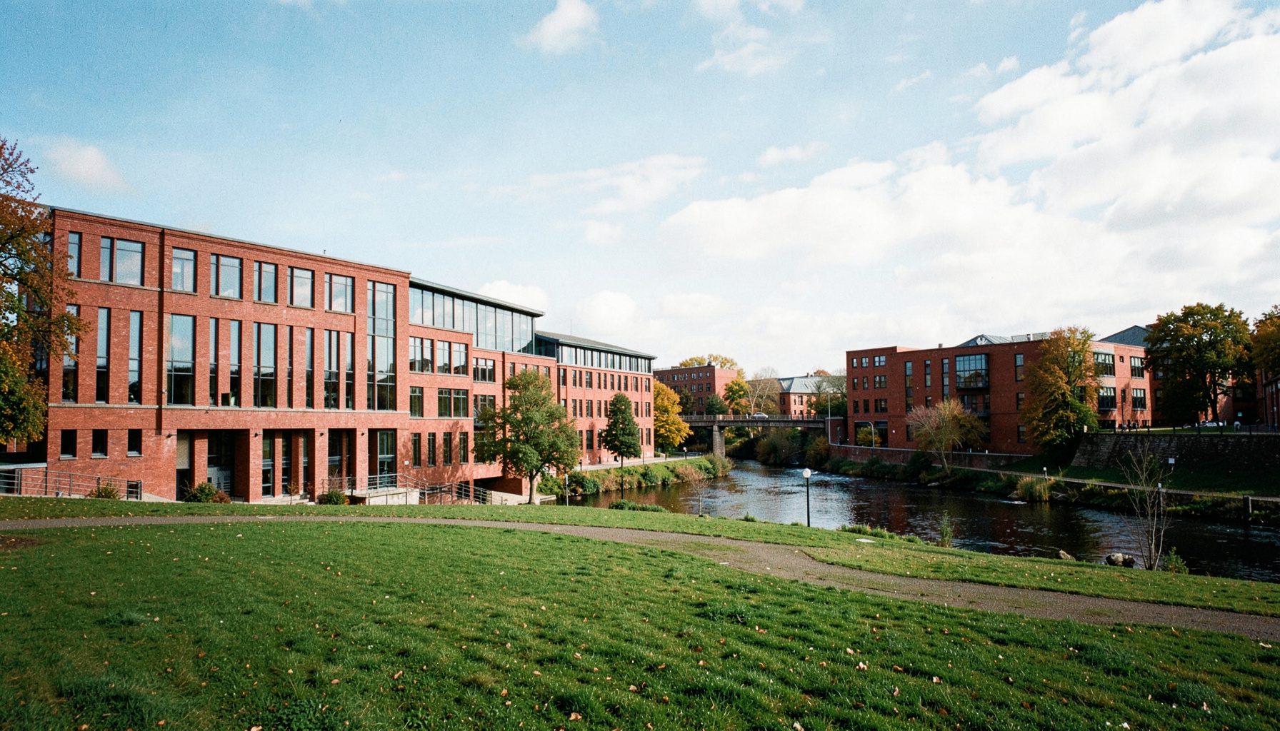 Wide-angle photograph of the University of Limerick campus on a bright day, showing the modern red-brick buildings along the River Shannon with green lawns and clear sky